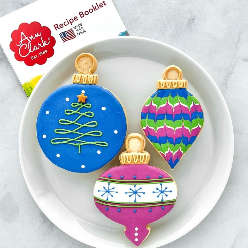 Three colorful, holiday dessert-themed ornament cookies rest on a marble surface, with a recipe booklet partially visible in the background.