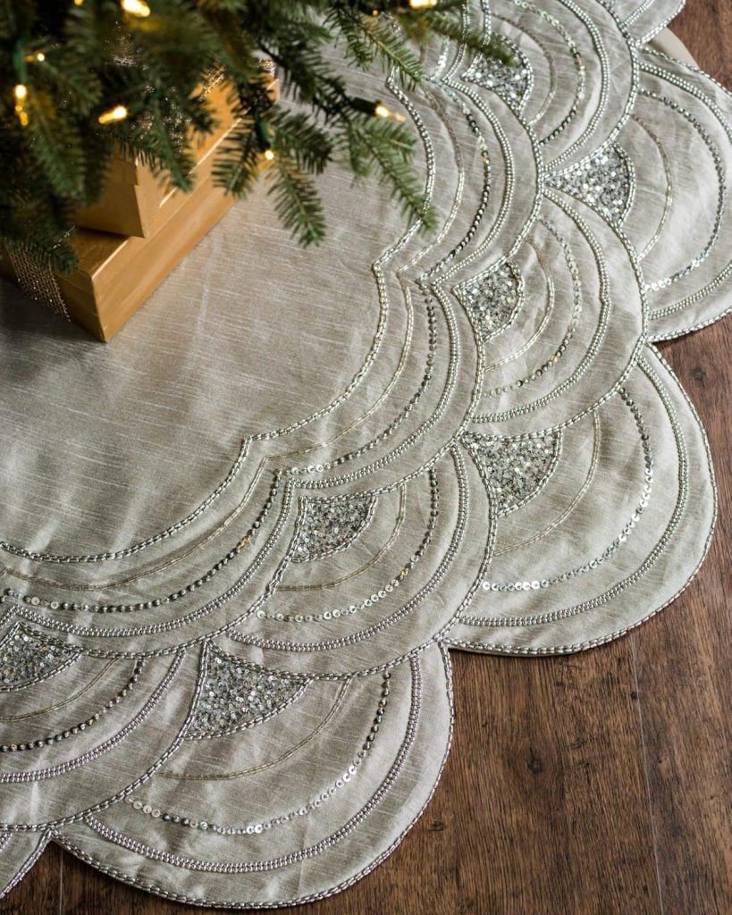 The elegant Christmas tree skirt, adorned with intricate beaded patterns, graces the wooden floor, partially covering the wrapped gifts.