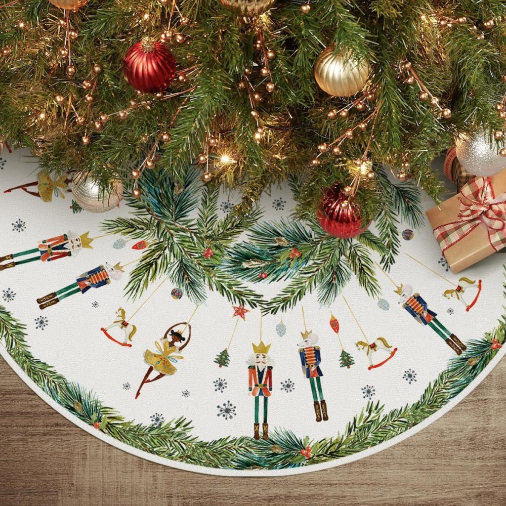 A Christmas tree with red and gold ornaments rests on a beautifully decorated Christmas tree skirt, adorned with nutcracker designs and green garland, surrounded by wrapped gifts on the wooden floor.