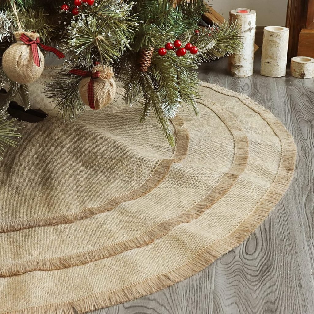 A charming burlap Christmas tree skirt nestles under a decorated tree adorned with red berries and burlap ornaments, set against the backdrop of a wooden floor with birch log candles softly glowing in the background.