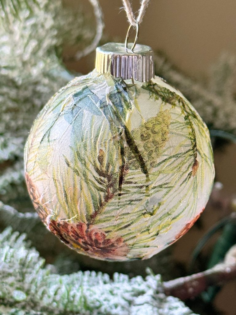 Close-up of a decorative glass ornament hanging on a frosted pine tree branch, featuring a design of pine needles and cones.