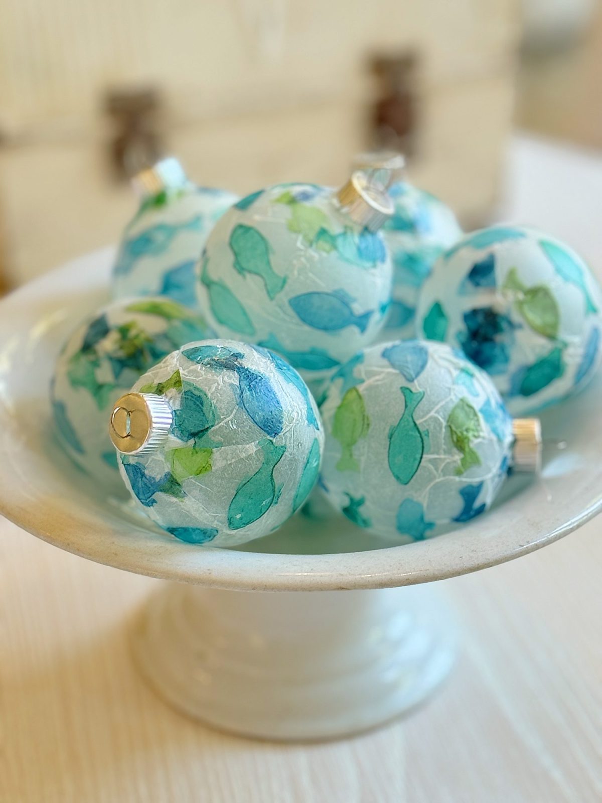 A white pedestal bowl holds several translucent ornaments with blue and green leaf patterns.