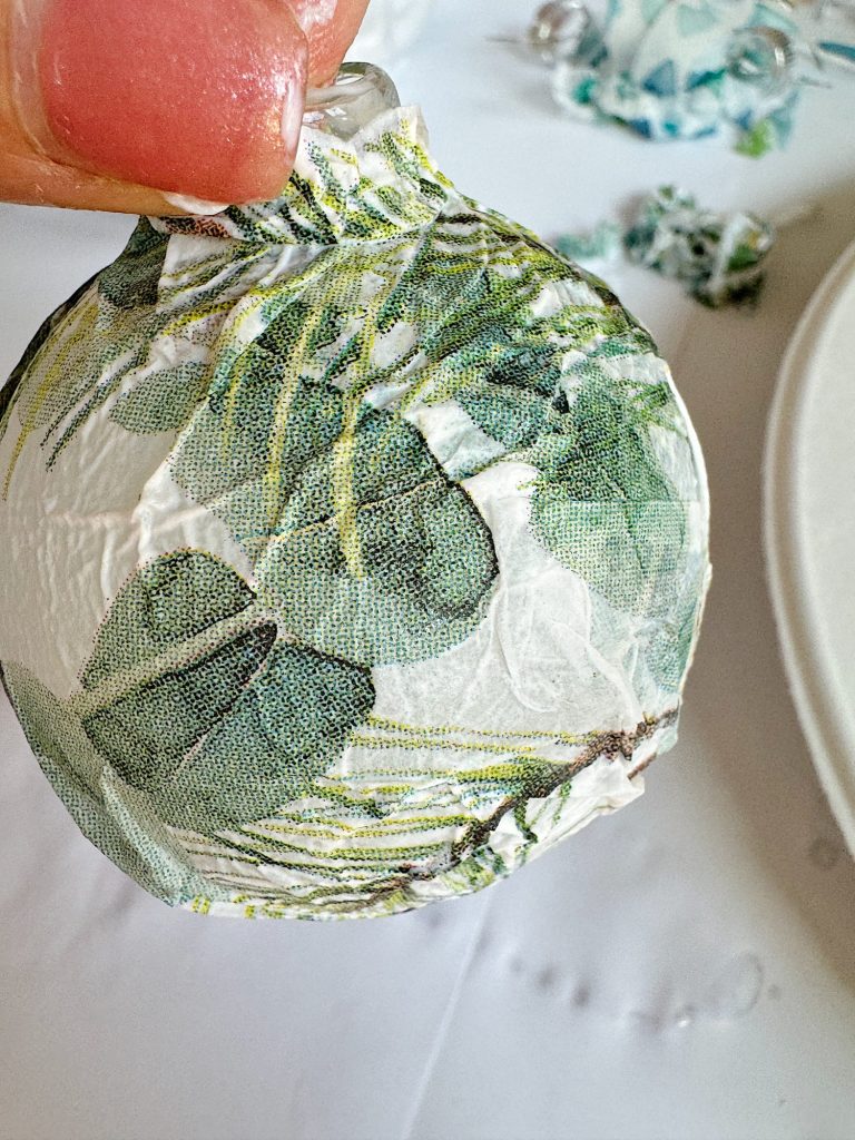 A hand holding a round ornament covered with decorative fabric featuring green leaves.