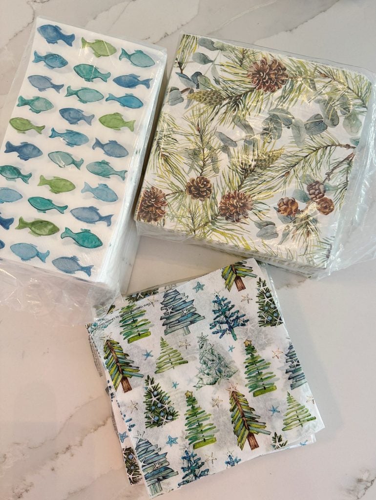 Three sets of decorative napkins are shown. One has a green fish pattern, another features pinecones and leaves, and the last has Christmas trees and stars.