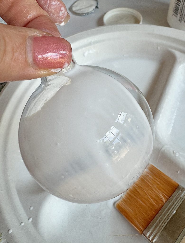 Hand holding a frosted glass ornament over a paint tray with a brush nearby.