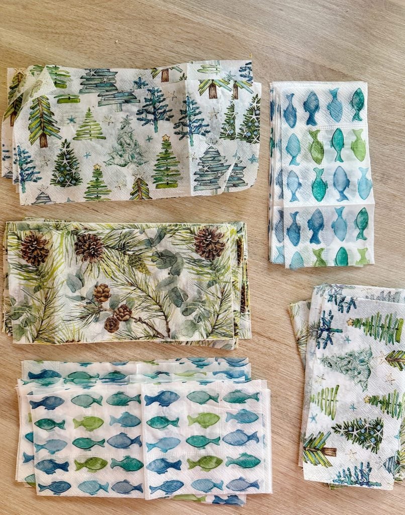 Five stacks of decorative napkins with various nature-themed designs, including trees and fish, are laid out on a wooden surface.