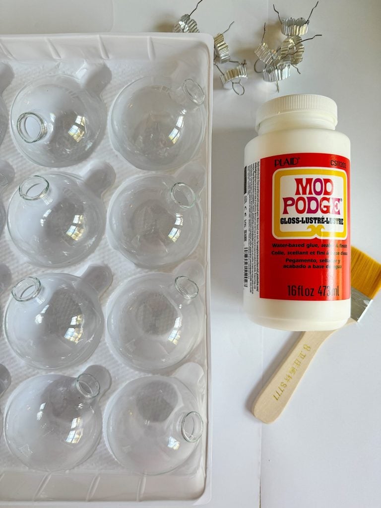 Clear plastic ornament balls in a tray, a bottle of Mod Podge, a paintbrush, and metal hooks are arranged on a white surface.