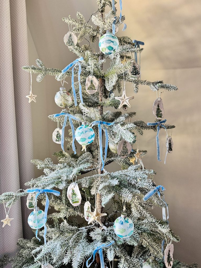 A frosted Christmas tree decorated with blue ribbons, round ornaments, and star-shaped decorations stands indoors.