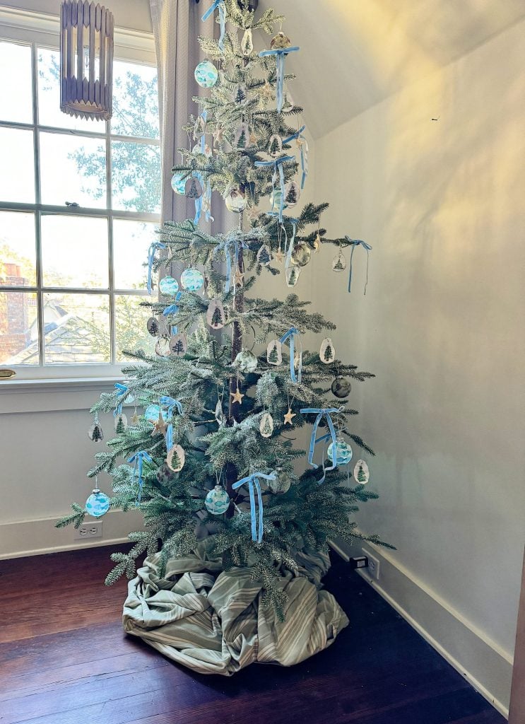 A tall Christmas tree adorned with blue and silver ornaments stands by a window. The base is wrapped in a striped fabric.