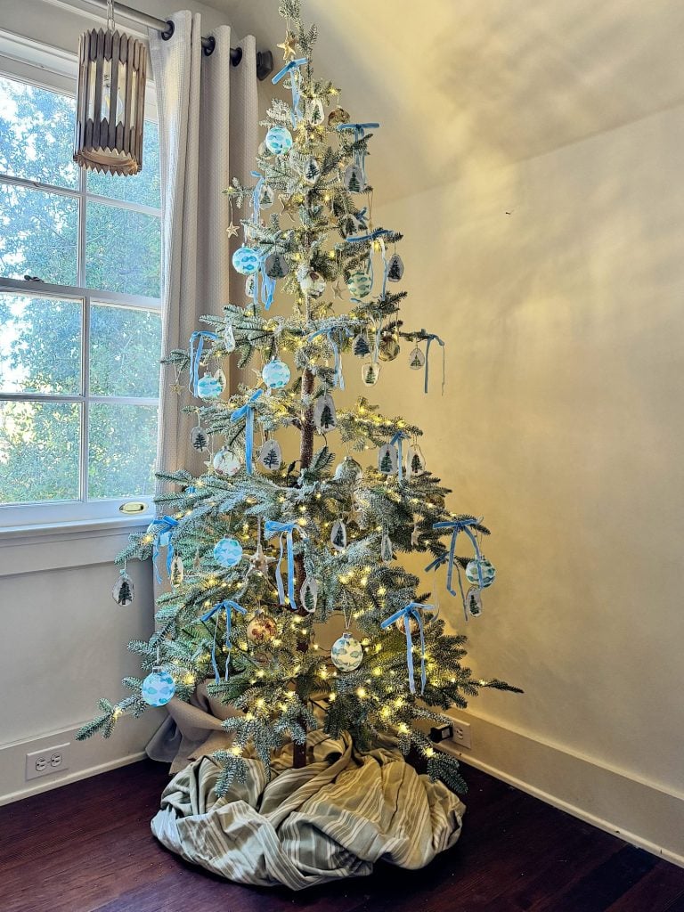 A decorated Christmas tree with blue and white ornaments and lights stands in a corner by a curtained window. The tree skirt is striped.