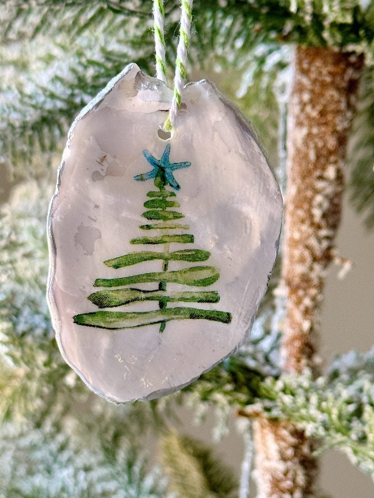 A handmade ornament with a painted green Christmas tree design hangs on a string, placed against a backdrop of a frosted tree branch.