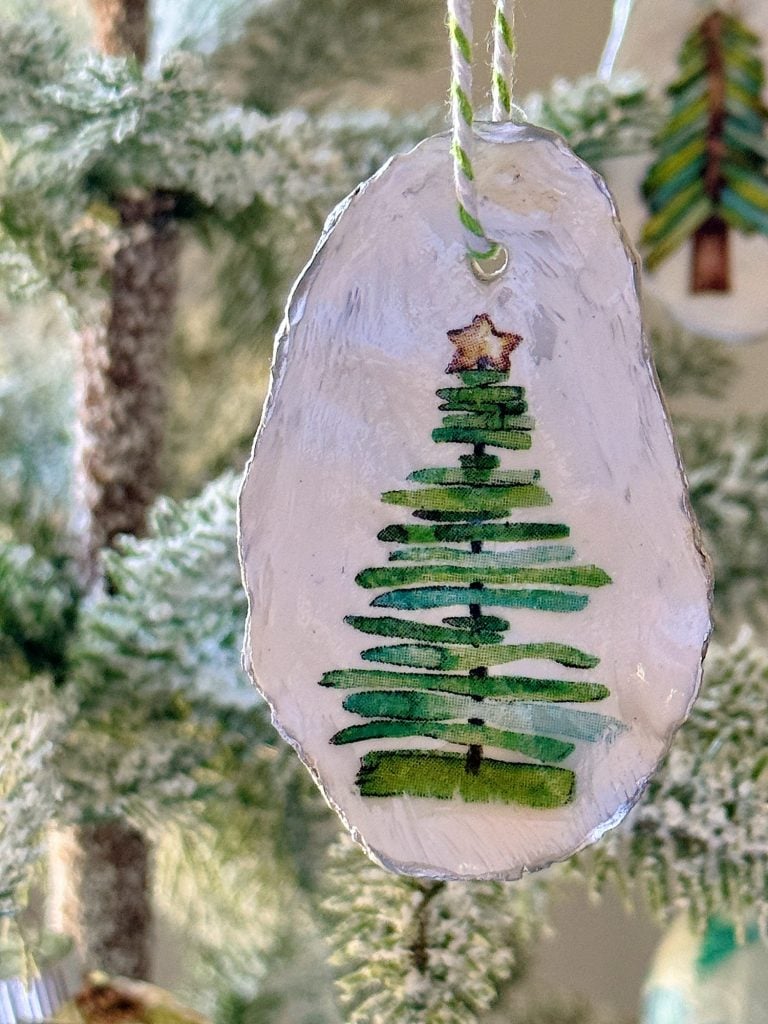 Close-up of a handmade Christmas ornament, painted with a green tree topped with a star, hanging on a snowy artificial tree branch.