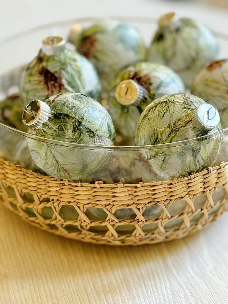 A woven basket holds several transparent glass ornaments with green foliage patterns, displayed on a light wooden surface.