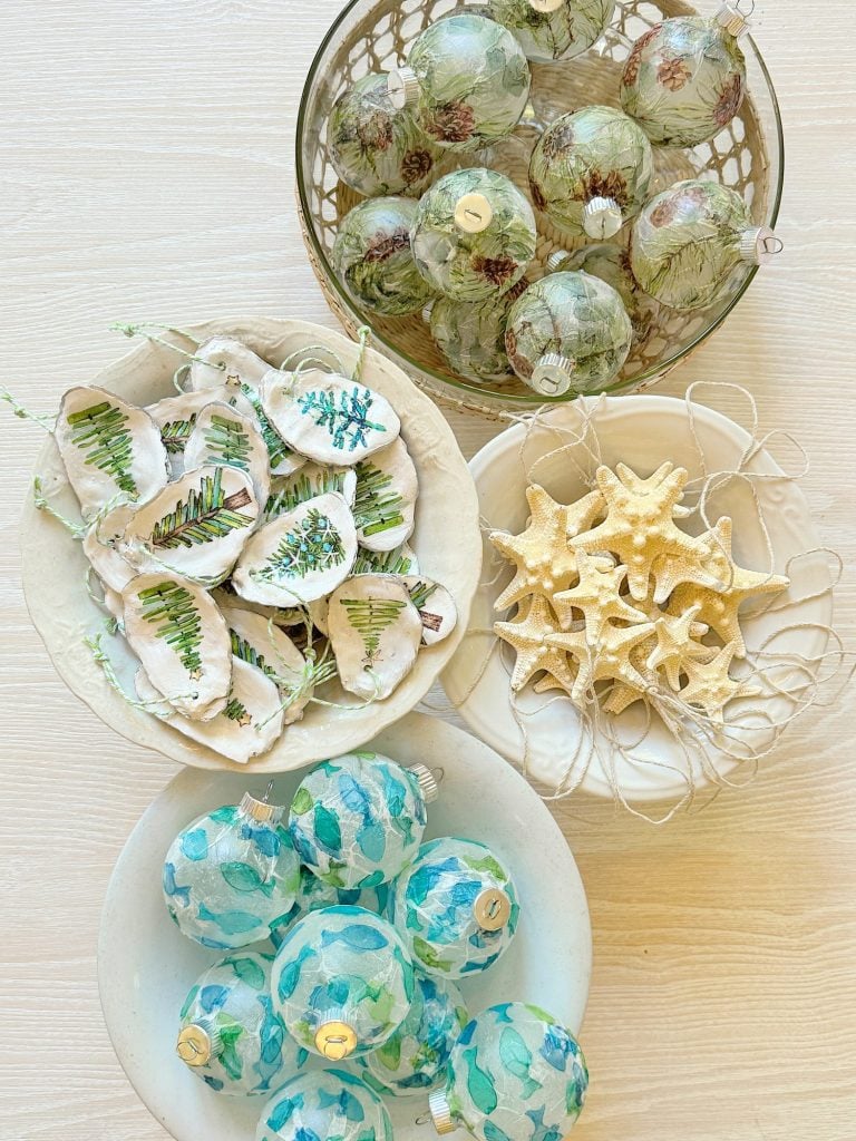 Bowls arranged with holiday decorations including green pinecone ornaments, leaf-patterned ornaments, white starfish, and blue assorted ornaments on a light wood surface.