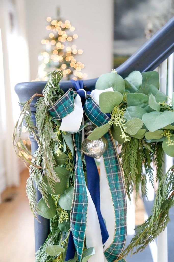 Festive staircase adorned with greenery, plaid ribbon, and silver ornaments in classic Christmas colors, with a blurred Christmas tree glowing warmly in the background.