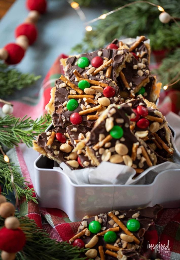 Festive chocolate bark with red and green candies, pretzels, and peanuts, displayed on a holiday-themed table with greenery and lights.