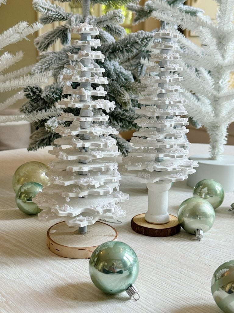 Two white ornamental trees surrounded by green metallic baubles on a light wooden surface.