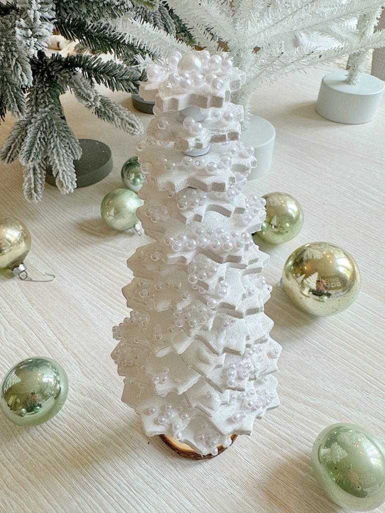 A cone-shaped white pastry decorated with star-shaped layers and small beads, surrounded by green and silver ornaments on a light wood surface.