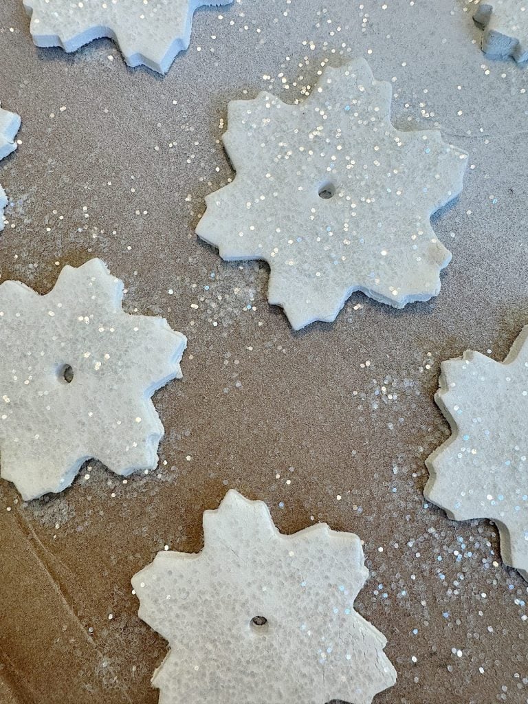 Snowflake-shaped cookies with a hole in the center are dusted with white sugar sprinkles on a brown baking sheet.