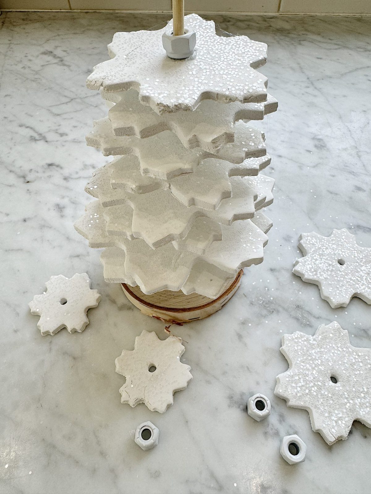 Stacked white snowflake-shaped ornaments with metal nuts on a marble surface. Some ornaments and nuts are scattered nearby.