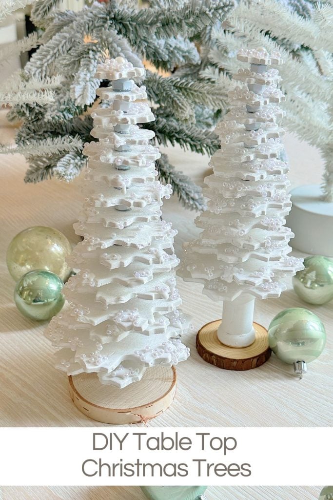 Two DIY tabletop Christmas trees made of stacked white scalloped layers with decorative white beads, set on a light wood surface with green ornaments and a frosted pine backdrop.