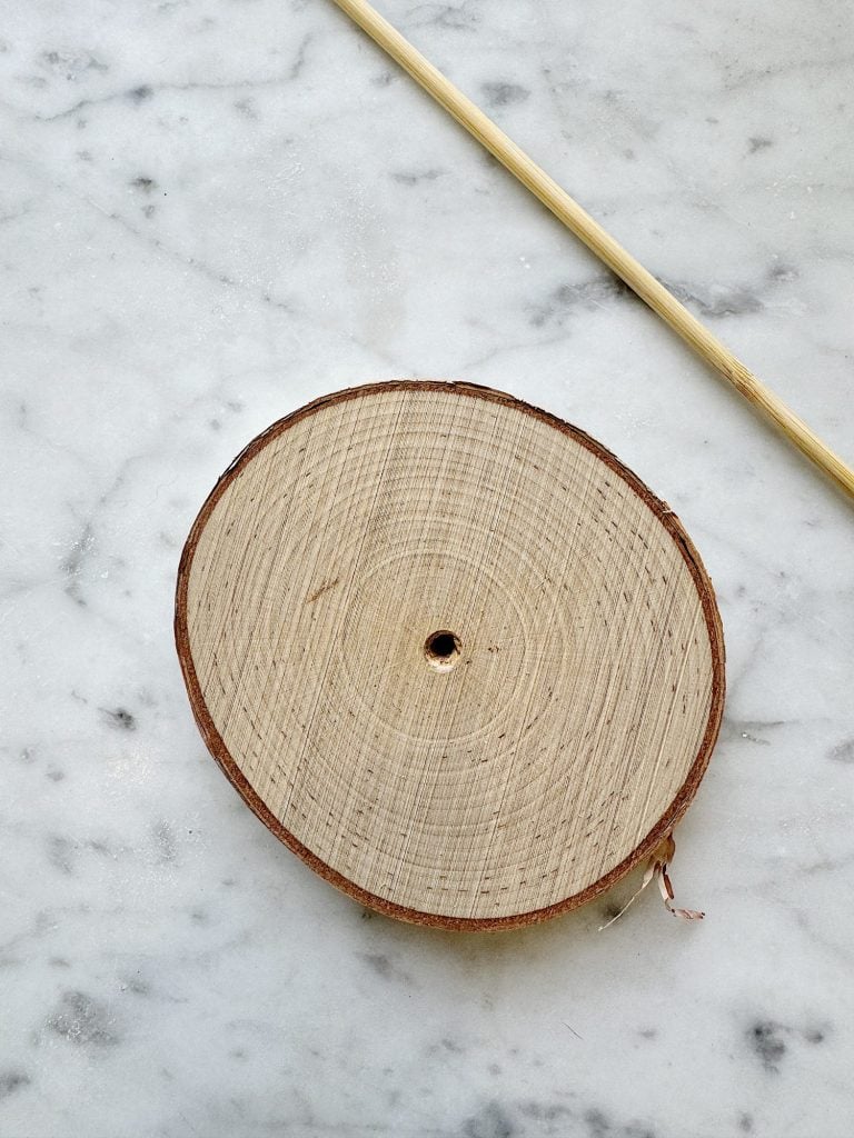 A round wooden slice with a small central hole lies on a marble surface next to a thin wooden stick.