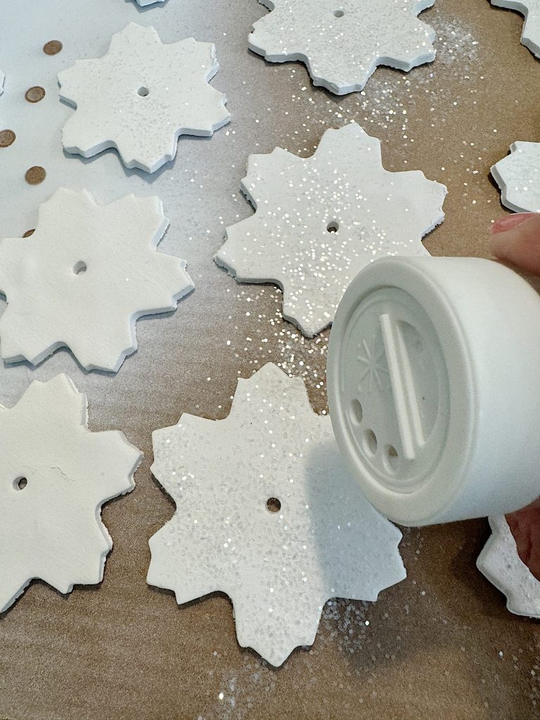 Hand sprinkling glitter on white clay snowflake-shaped ornaments on a table.