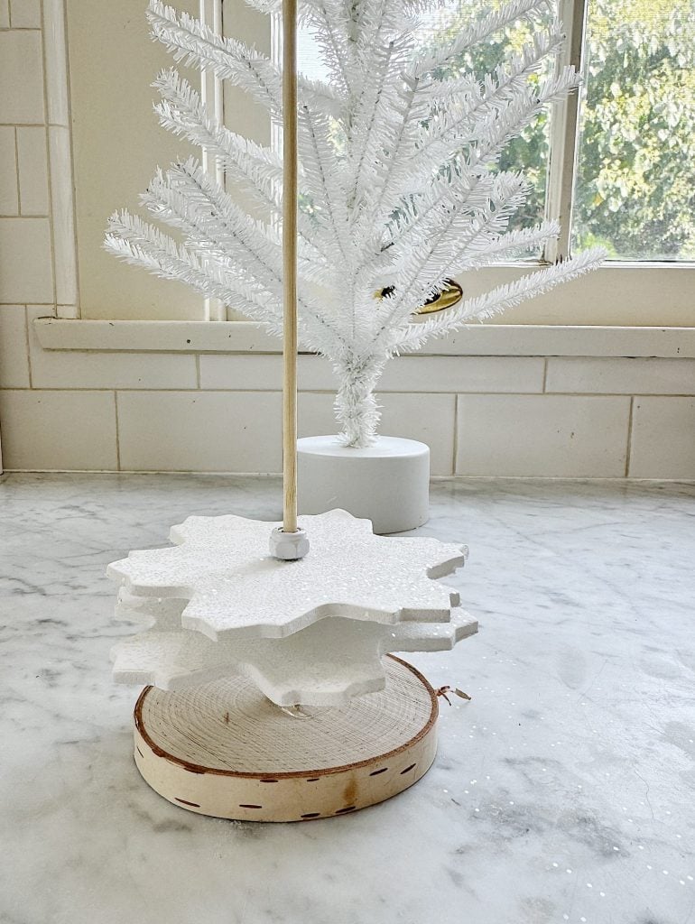 White decorative snowflake ornament on a wooden base in front of a tabletop white tree, set on a marble surface near a window.