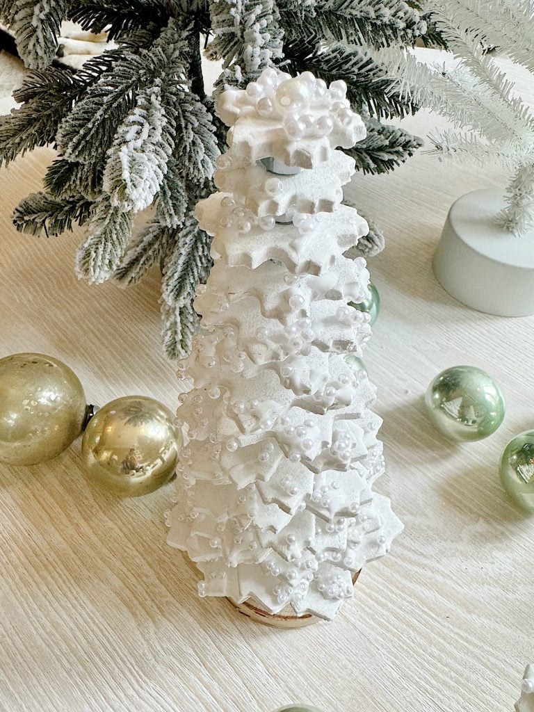 A decorative, white, star-shaped cookie tower resembling a Christmas tree, adorned with white pearls and frosting, is placed on a wooden surface with frosted pine branches and gold ornaments nearby.