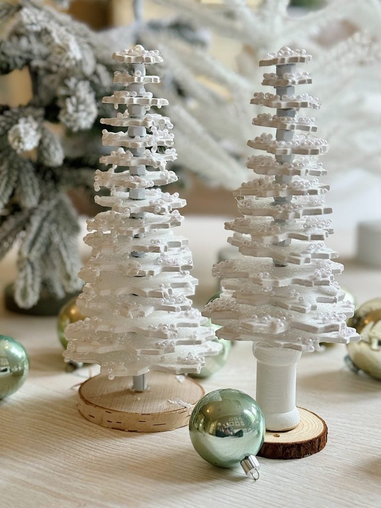 Two white miniature Christmas trees made of stacked layers stand on a wooden surface, with green ornaments and frosted pine branches in the background.