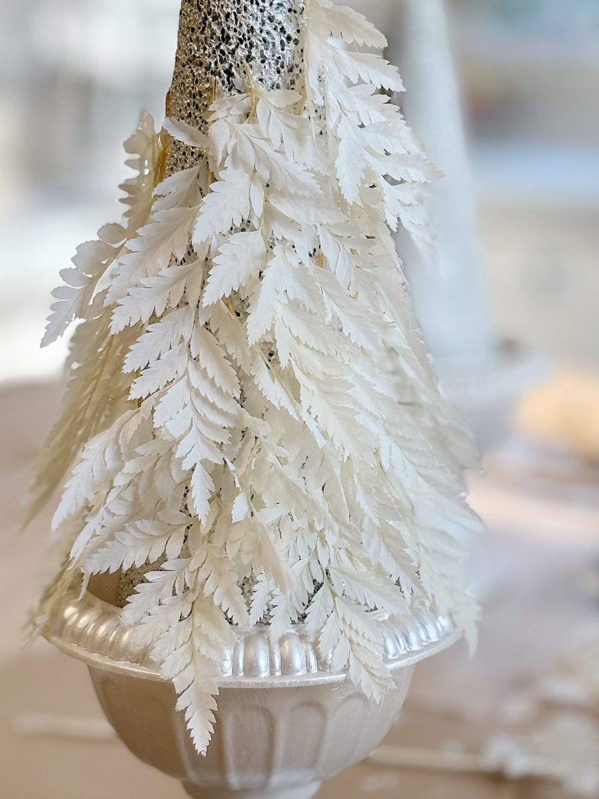 A decorative arrangement of white, leafy branches is placed in a white, textured vase with a blurred background.
