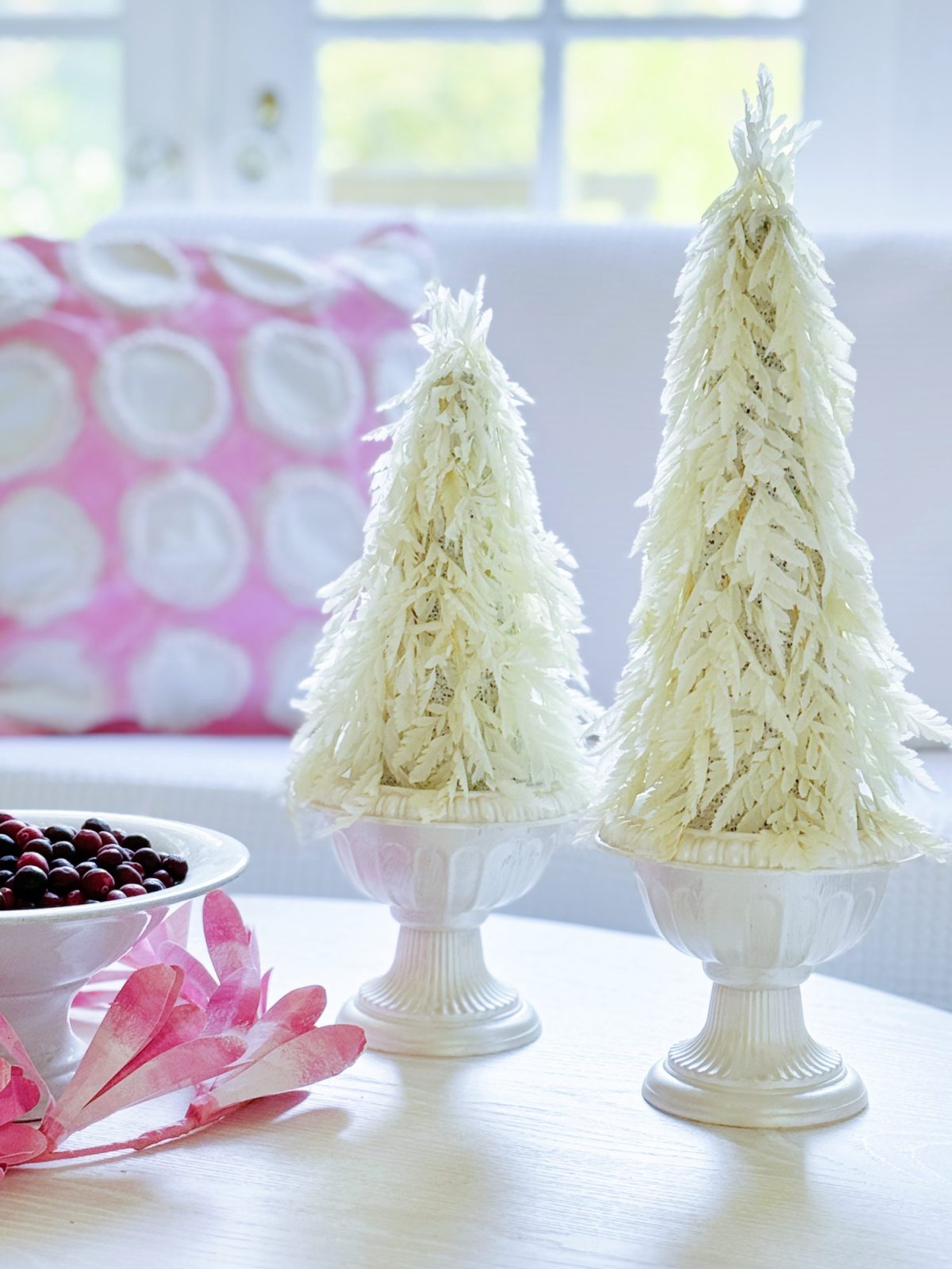 Two decorative white Christmas trees in ceramic vases on a table with a bowl of red berries and a pink pillow with white circles in the background.