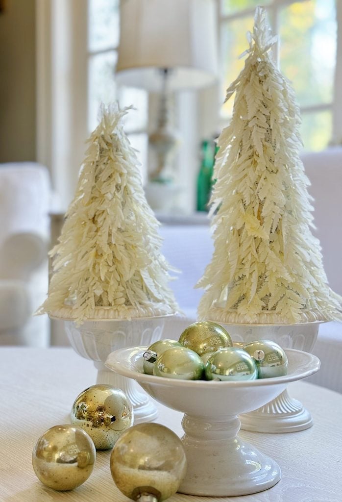 Two decorative white Christmas trees in pots on a table with gold ornaments in a dish and scattered nearby.