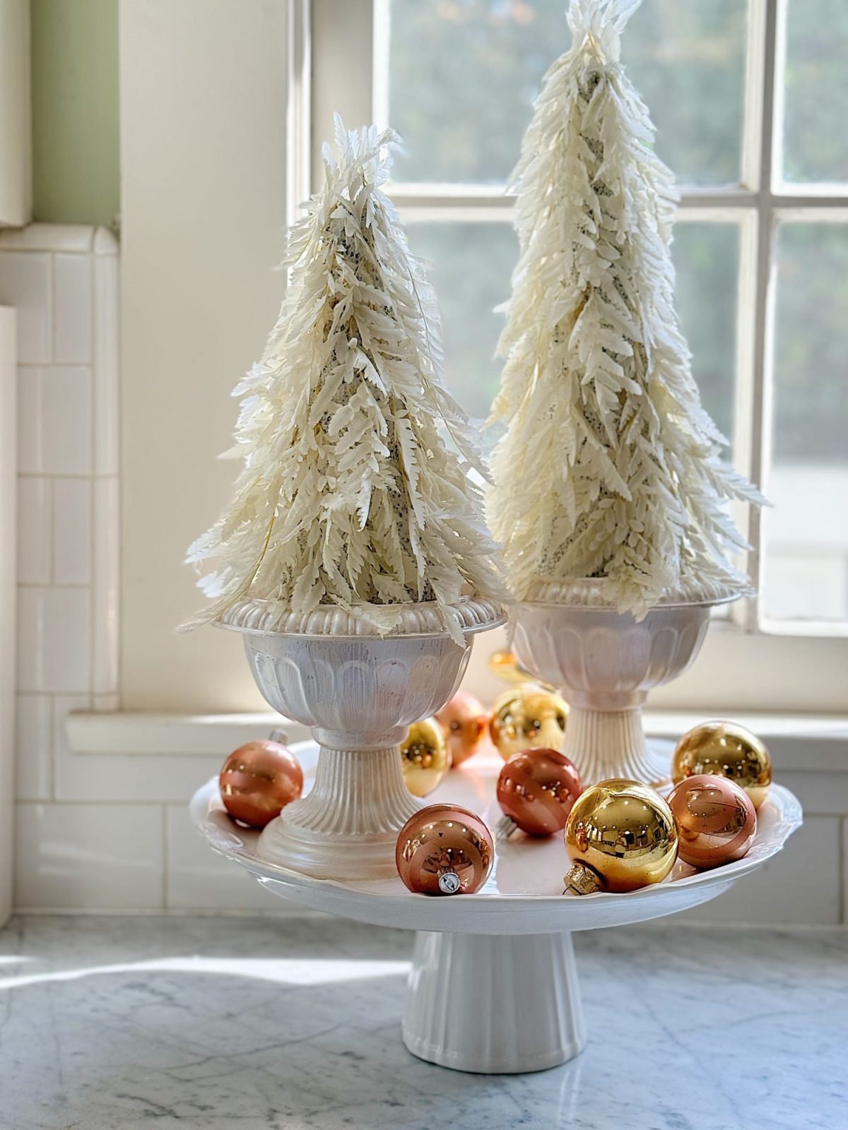 Two white decorative trees in vases on a pedestal, surrounded by gold ornaments, are displayed in front of a window.