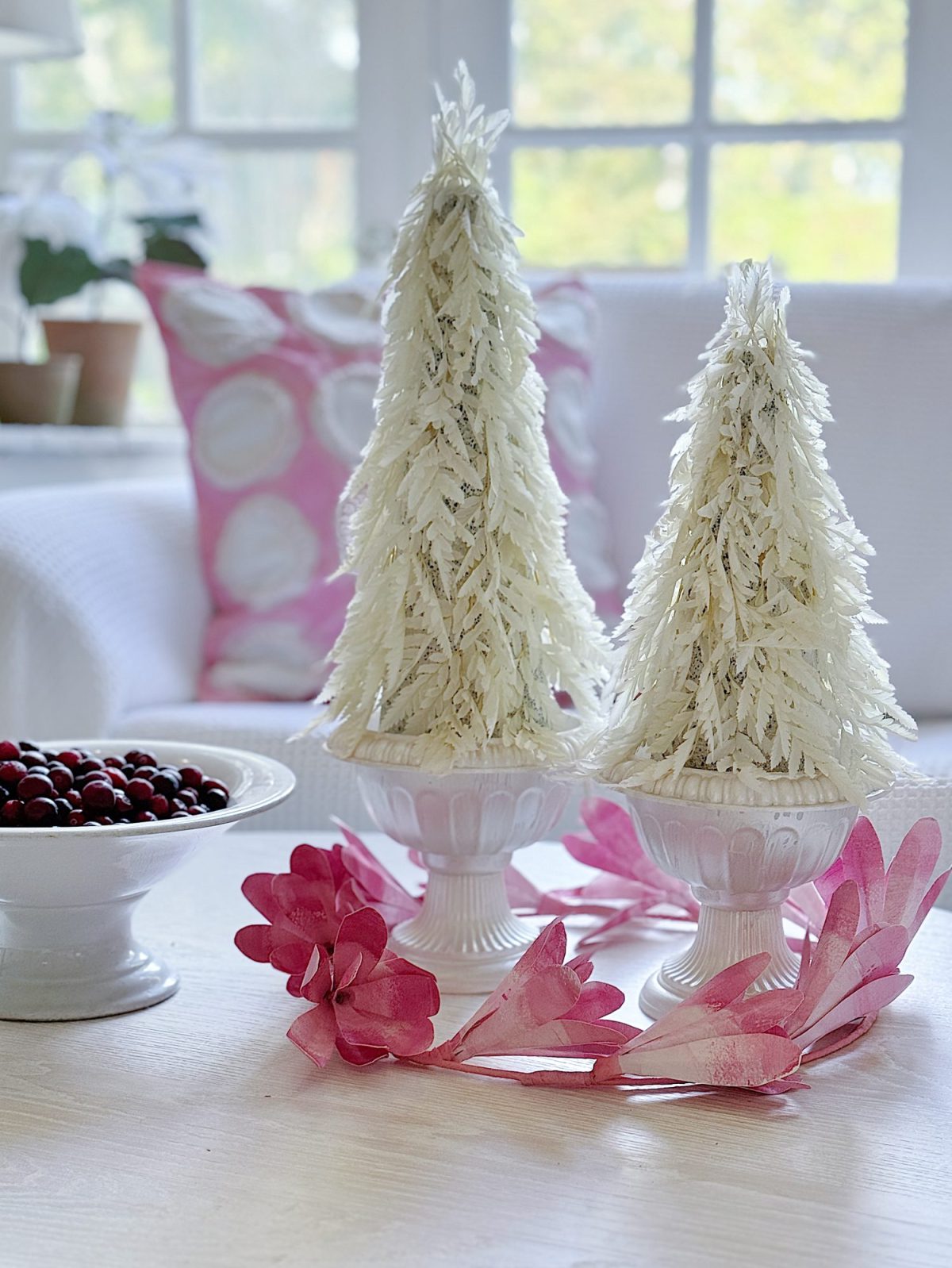 Two white decorative trees in vases on a table with a bowl of cranberries and pink ornaments, set against a bright window background.