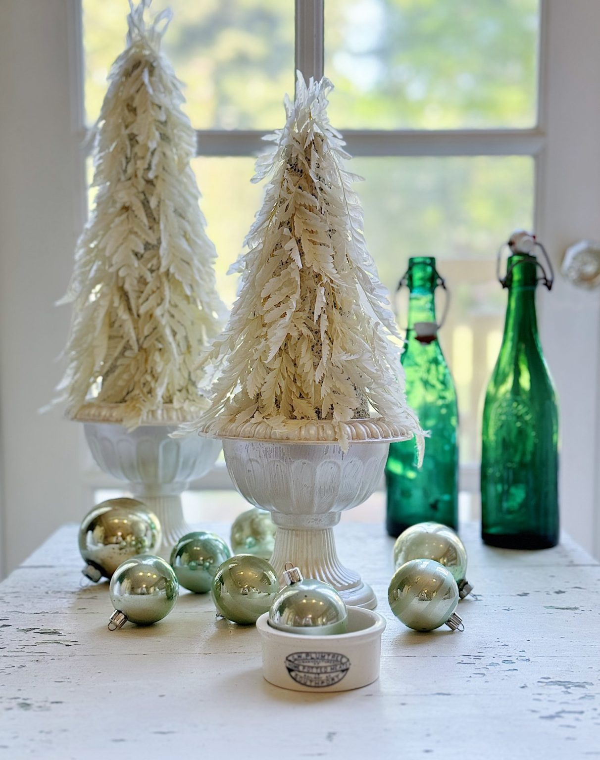 Two white feather trees in pots on a table with green vintage bottles and green-gold ornaments. Background shows a window with light.