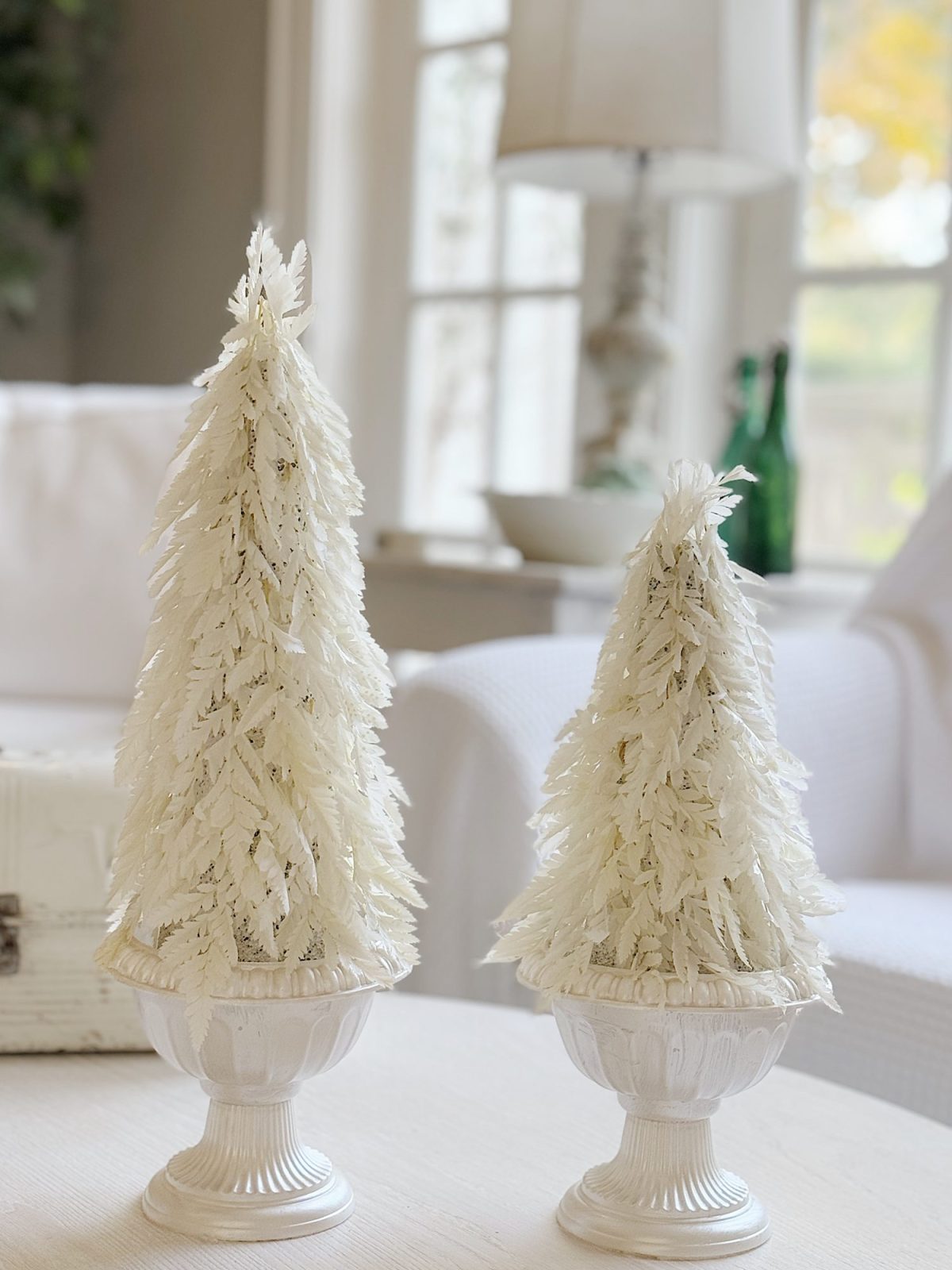 Two decorative white feather trees in ornate pots on a table in a bright room with a couch and lamp in the background.