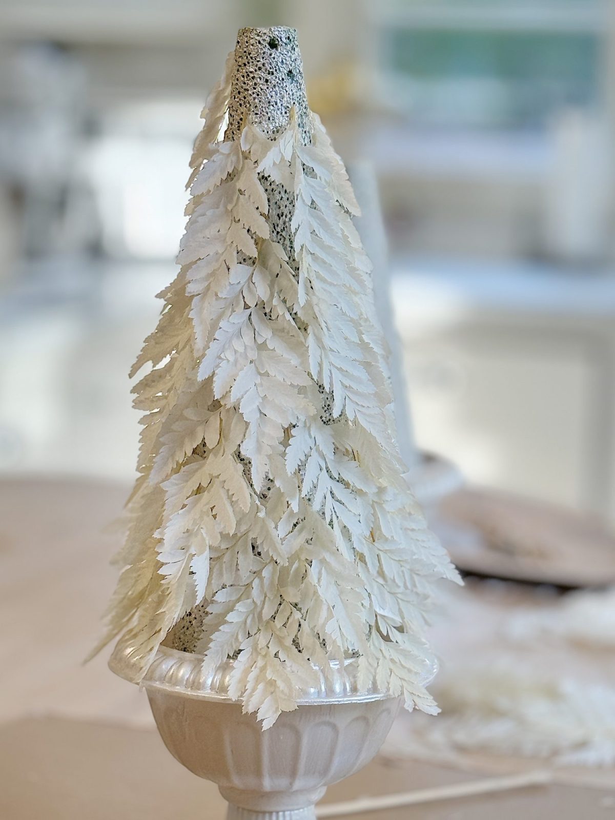A decorative object resembling a pine tree, made of layered white leaves, placed in a white pot on a light-colored surface.