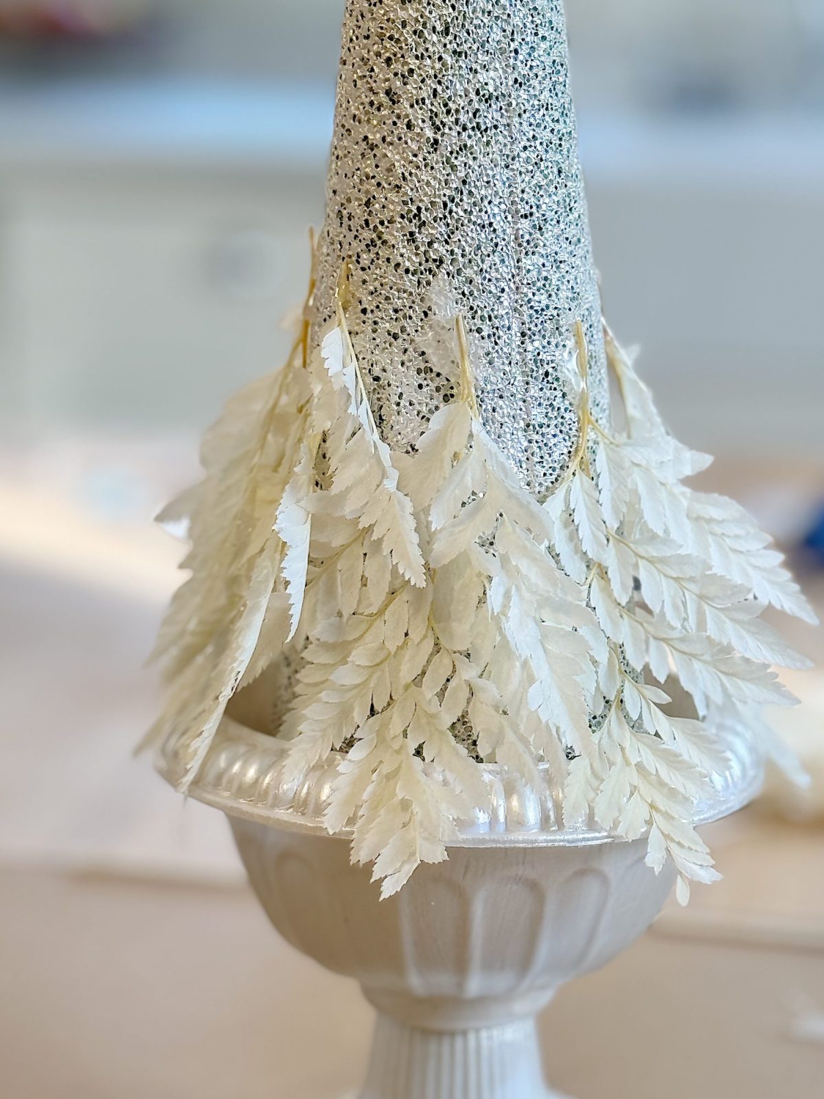A decorative cone with white glitter and leaf-like accents stands in a white urn-shaped base.