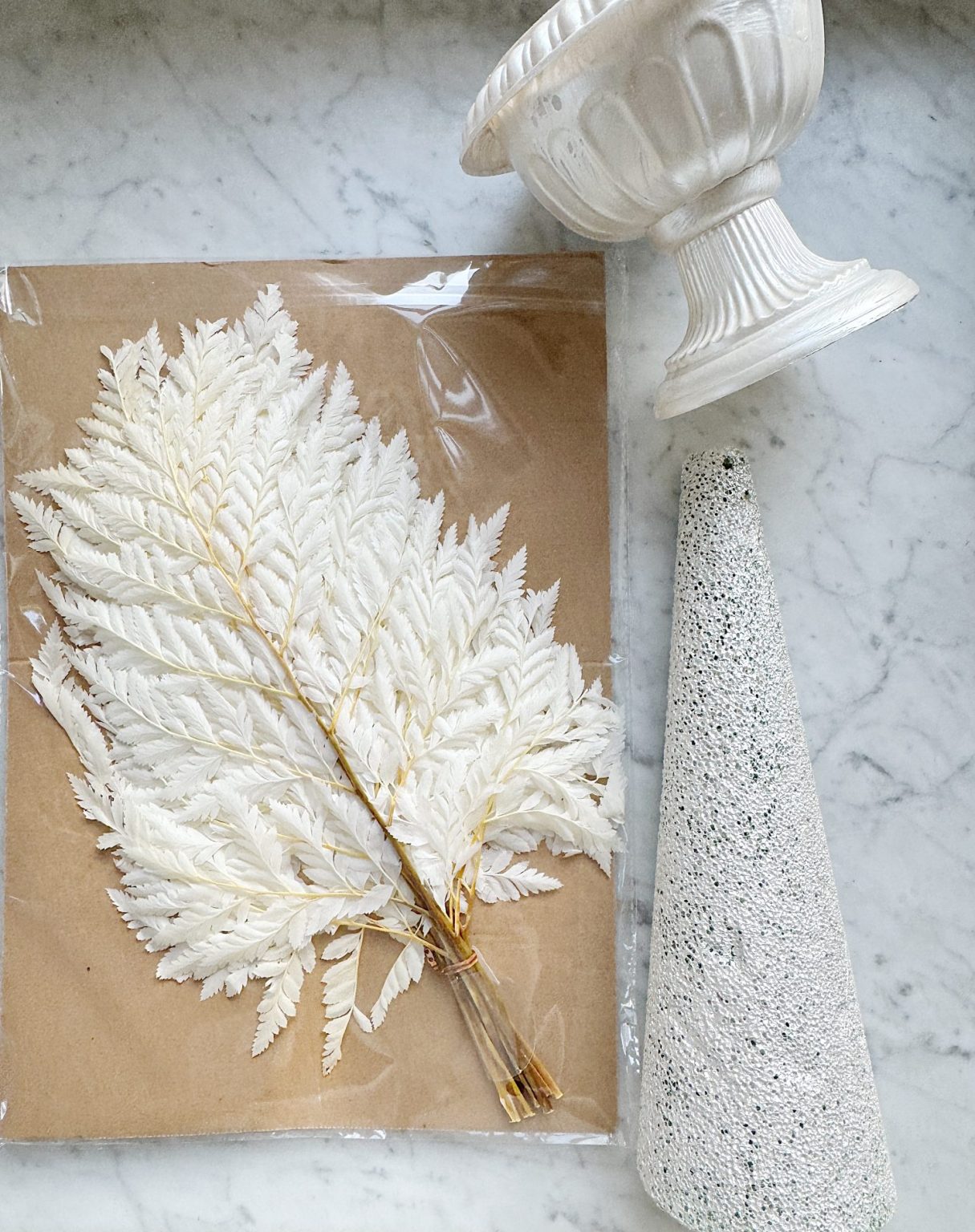 A bundle of white ferns, a white ceramic vase, and a textured white cone are arranged on a marble surface.