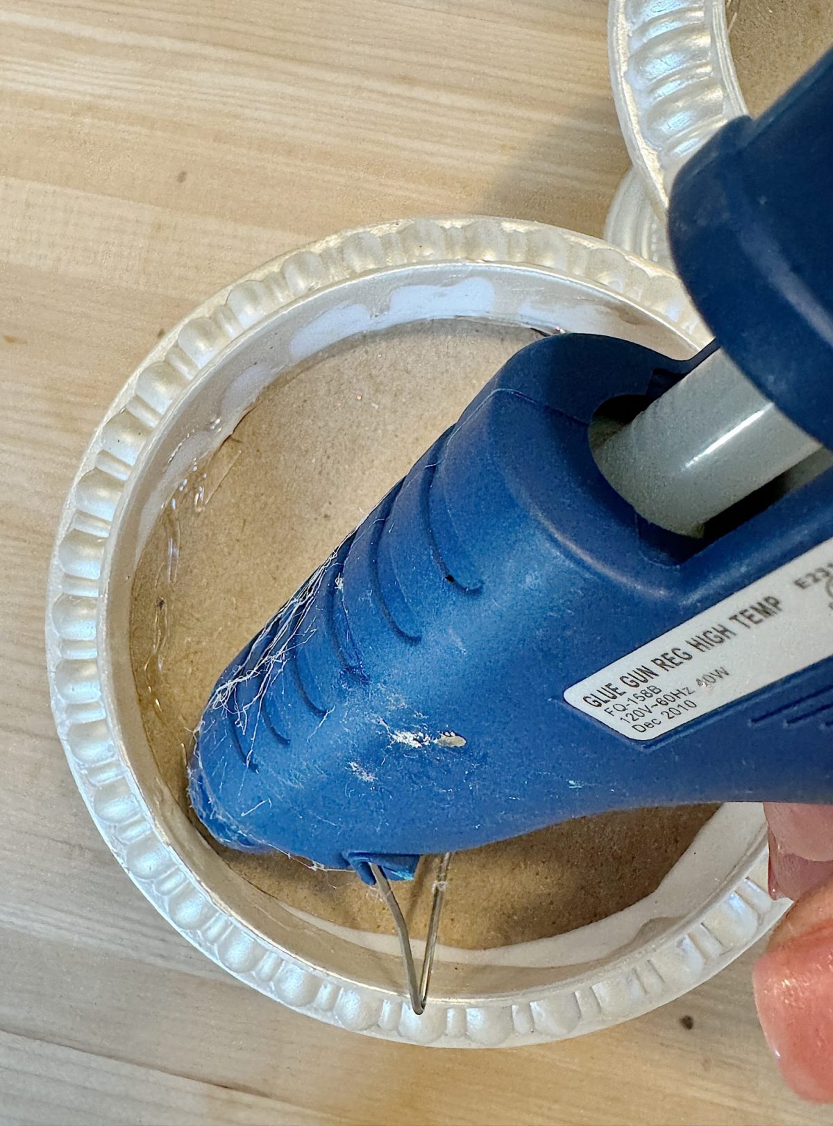 Close-up of a blue hot glue gun applying glue to a small round cardboard inside a white textured container on a wooden surface.