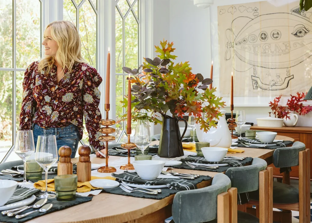 A woman stands by a dining table set with autumn-themed decor, featuring candles, leafy centerpieces, and place settings. Large windows provide natural light in the background.