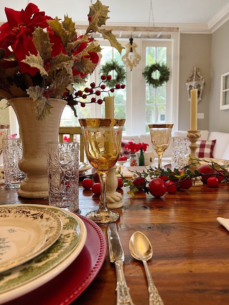 Festive dining table setting with gold-rimmed glasses, elegant plates, silverware, red and green decor, and candles. Holiday wreaths adorn the windows in the background, setting a perfect scene for Black Friday gatherings.