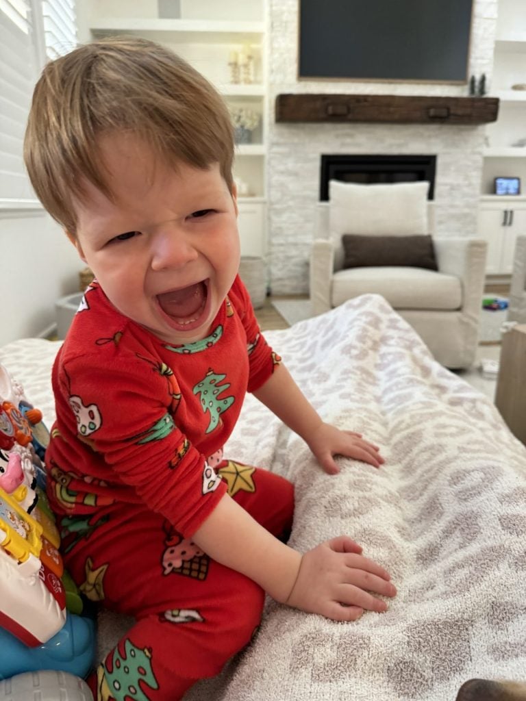 A young child in red pajamas is laughing while sitting on a couch in a cozy living room.