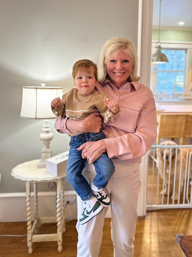 A woman in a pink shirt holds a child in a beige sweater. They stand in a living room with a lamp, a small table, and a glimpse into a kitchen area.