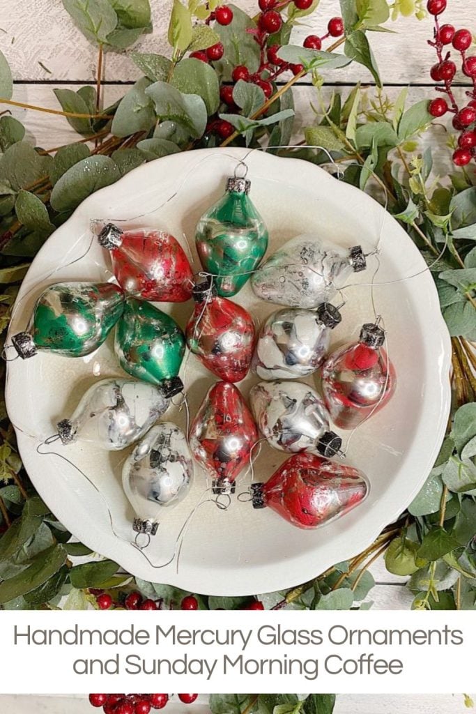 A white bowl with red, green, and silver Christmas ornaments, surrounded by green leaves and red berries on a wooden surface.