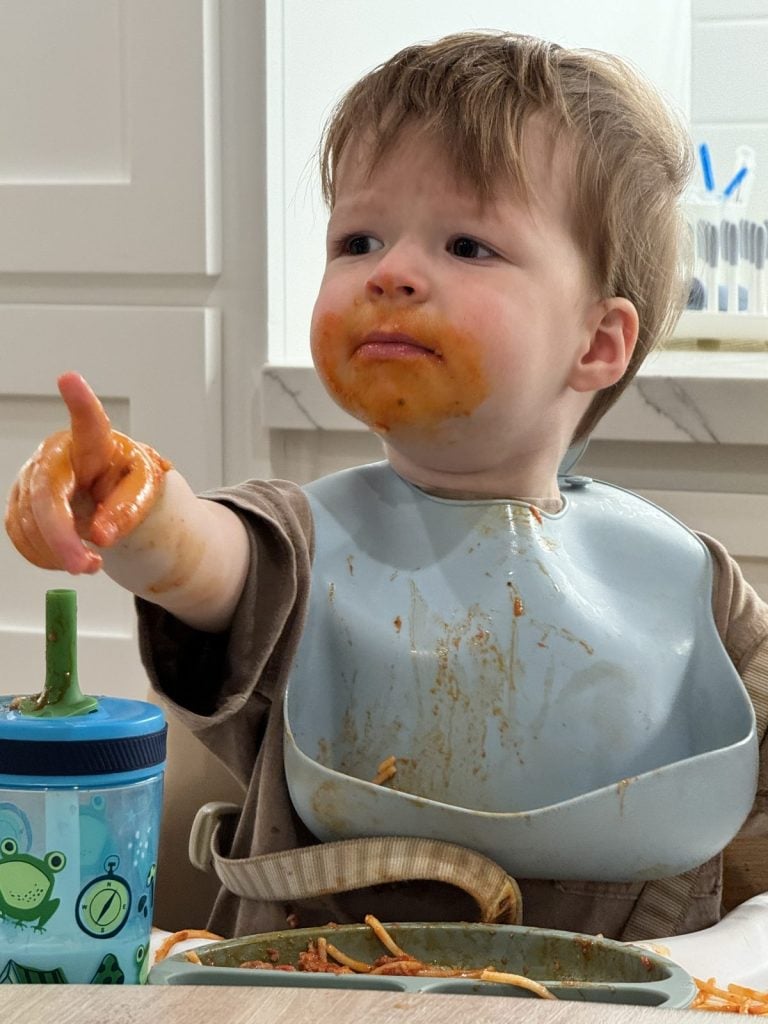 A toddler with a messy face points at something exciting, possibly the best Black Friday deal ever, while eating spaghetti. A sippy cup is on the table.