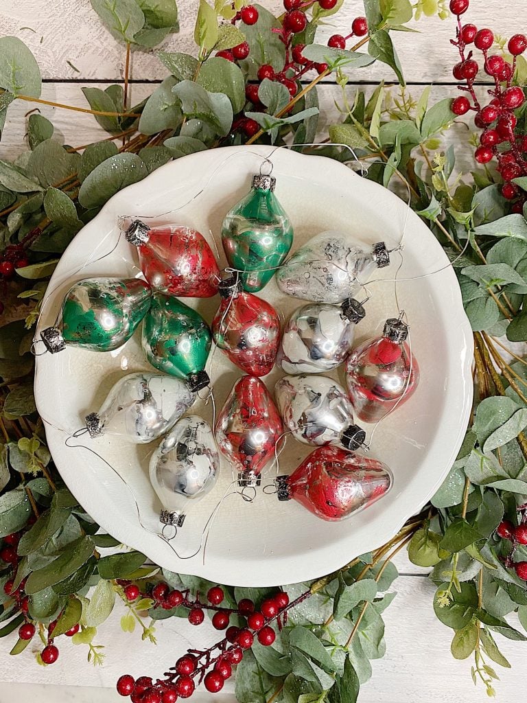 A white bowl with red, green, and silver Christmas ornaments, surrounded by green leaves and red berries on a wooden surface.