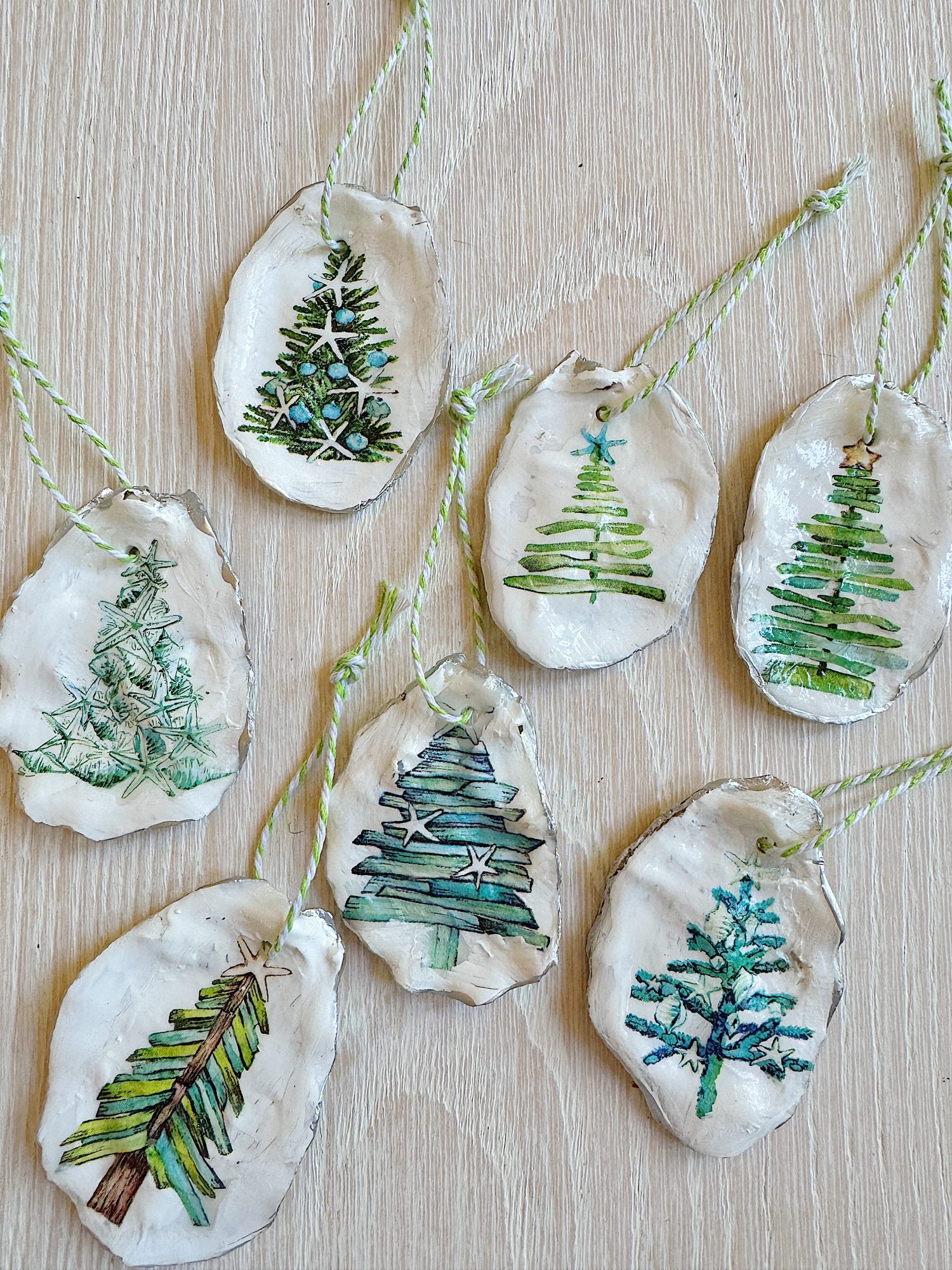 Seven handmade clay ornaments with painted Christmas tree designs, each hanging from green and white twine on a wooden surface.