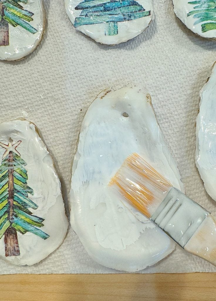 A paintbrush applies a white coat onto a tree-shaped ornament placed on a paper towel, next to other painted ornaments.